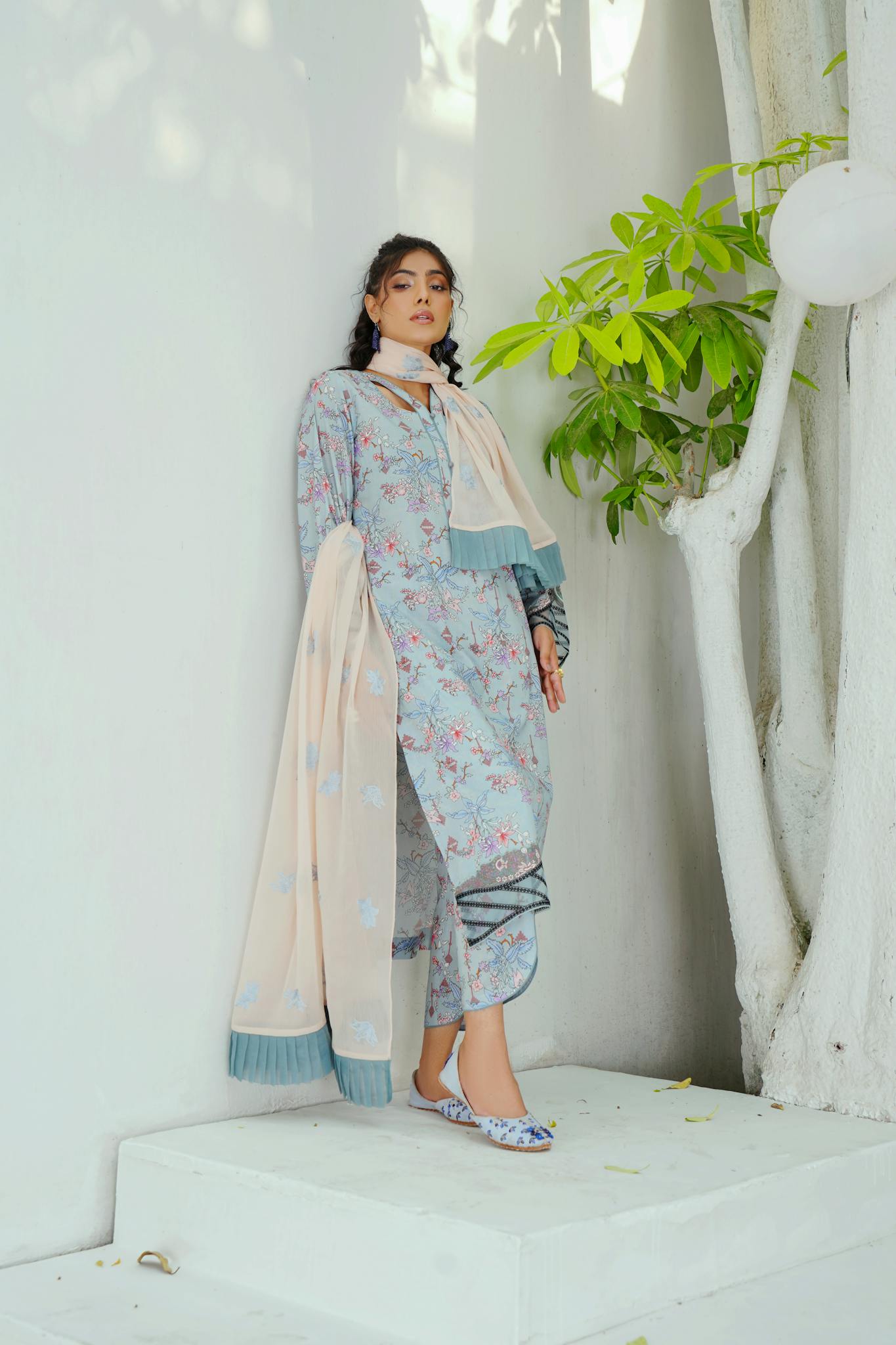 A woman in a traditional floral gown and scarf standing indoors with minimalistic white backdrop